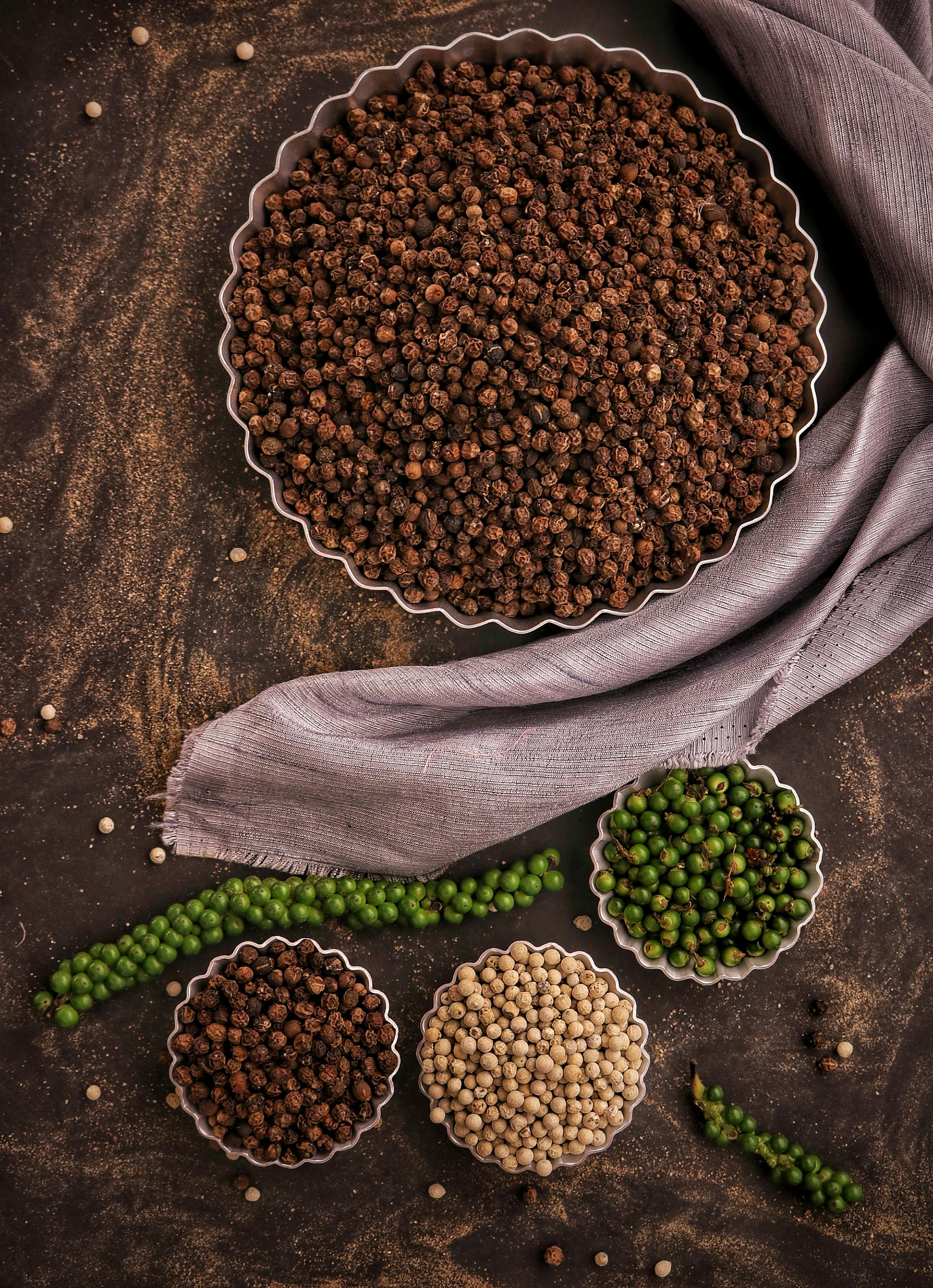 A variety of peppercorns displayed in bowls on a textured surface with a cloth.