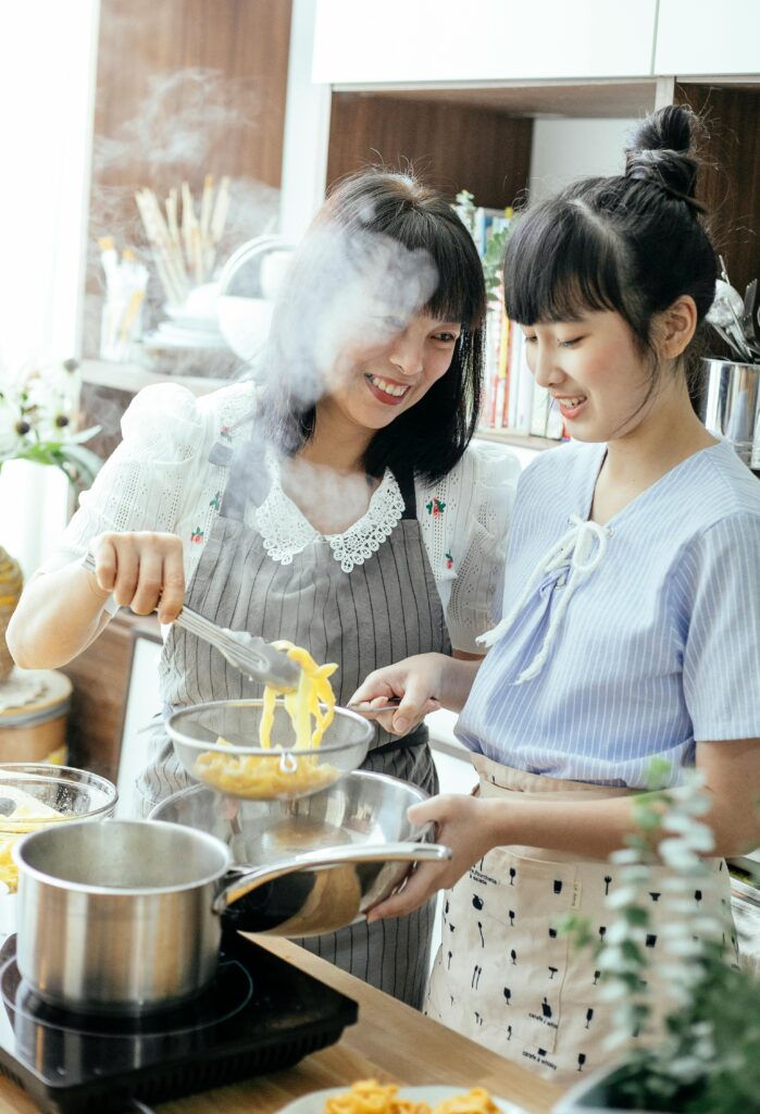 Happy young ethnic female with teenage daughter in aprons cooking homemade noodles and smiling while preparing dinner together in kitchen