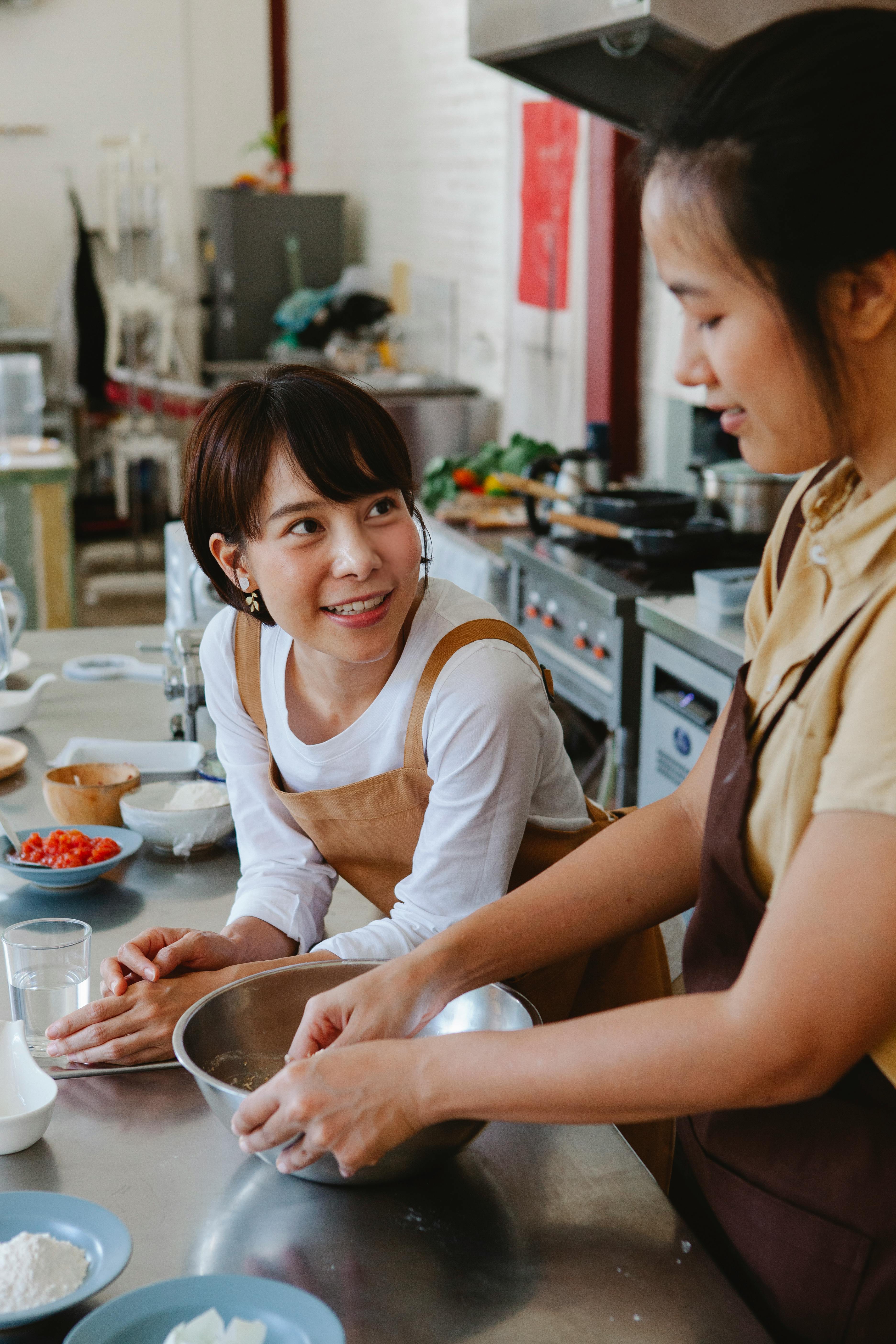 Two Asian women engaging in cooking, sharing smiles and tasks in a contemporary kitchen setting.